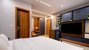 A modern hotel room with a large bed, a TV mounted on a dark panel, a small desk, and a seating area with a chair and table. - Imperial Stayz (Gurugram)