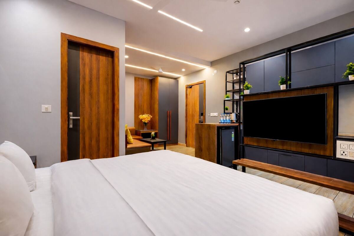 A modern hotel room with a large bed, a TV mounted on a dark panel, a small desk, and a seating area with a chair and table.