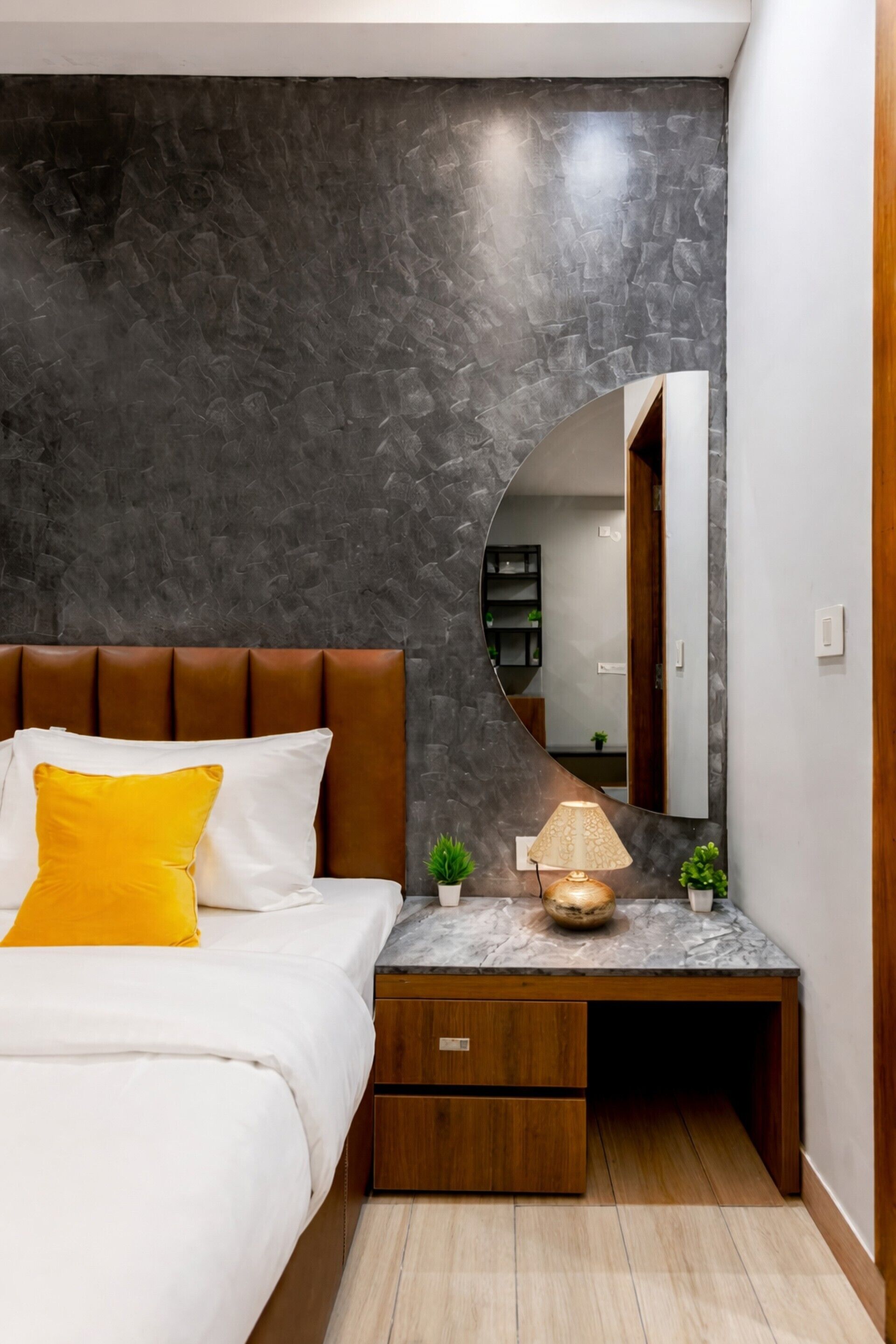 A bedroom with a curved wall, a bed with white and yellow pillows, a bedside table with a lamp, and a mirror.