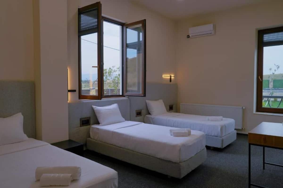A hotel room with two single beds, a small table, and a window with blinds.