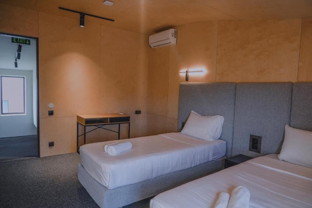 Habitación de hotel con dos camas, un escritorio, una silla, un equipo de aire acondicionado y una señal de salida.