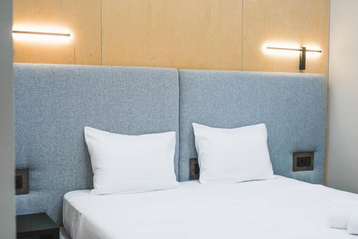 A hotel room with a bed, two white pillows, a grey headboard, and wall-mounted lighting.