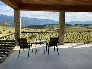 Een overdekt terras met een tafel en stoelen, stenen zuilen en uitzicht op een bergachtig landschap.