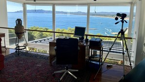 A room with a desk, computer, and telescope, overlooking a coastal view.