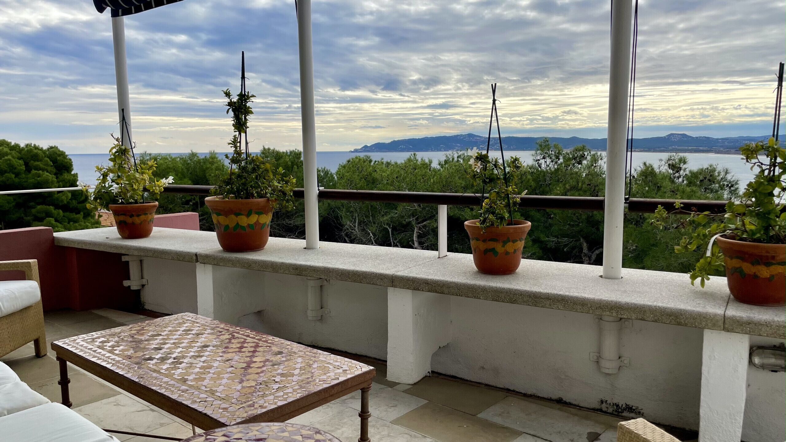 Varanda com plantas em vasos, mesa de azulejos e vista para o mar e montanhas.