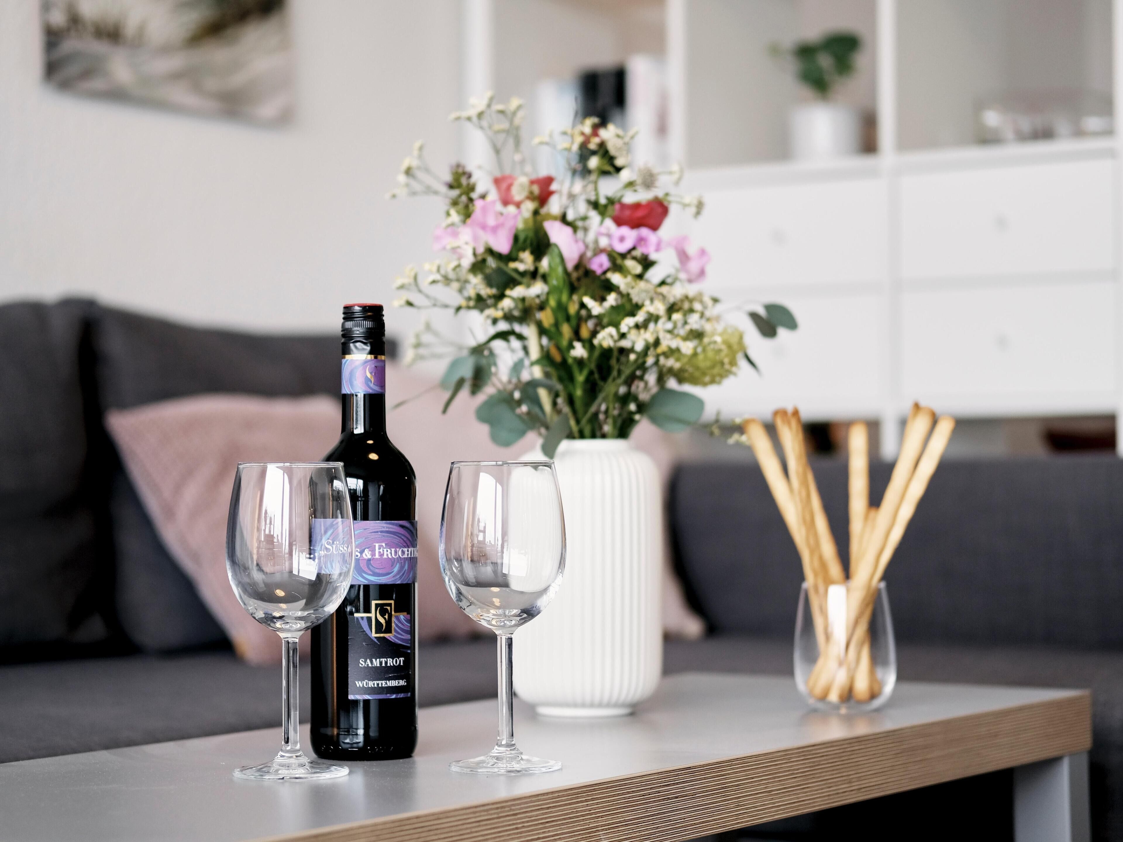 Eine Weinflasche, zwei Gläser, eine Vase mit Blumen und eine Schale mit Grissini auf einem Tisch.