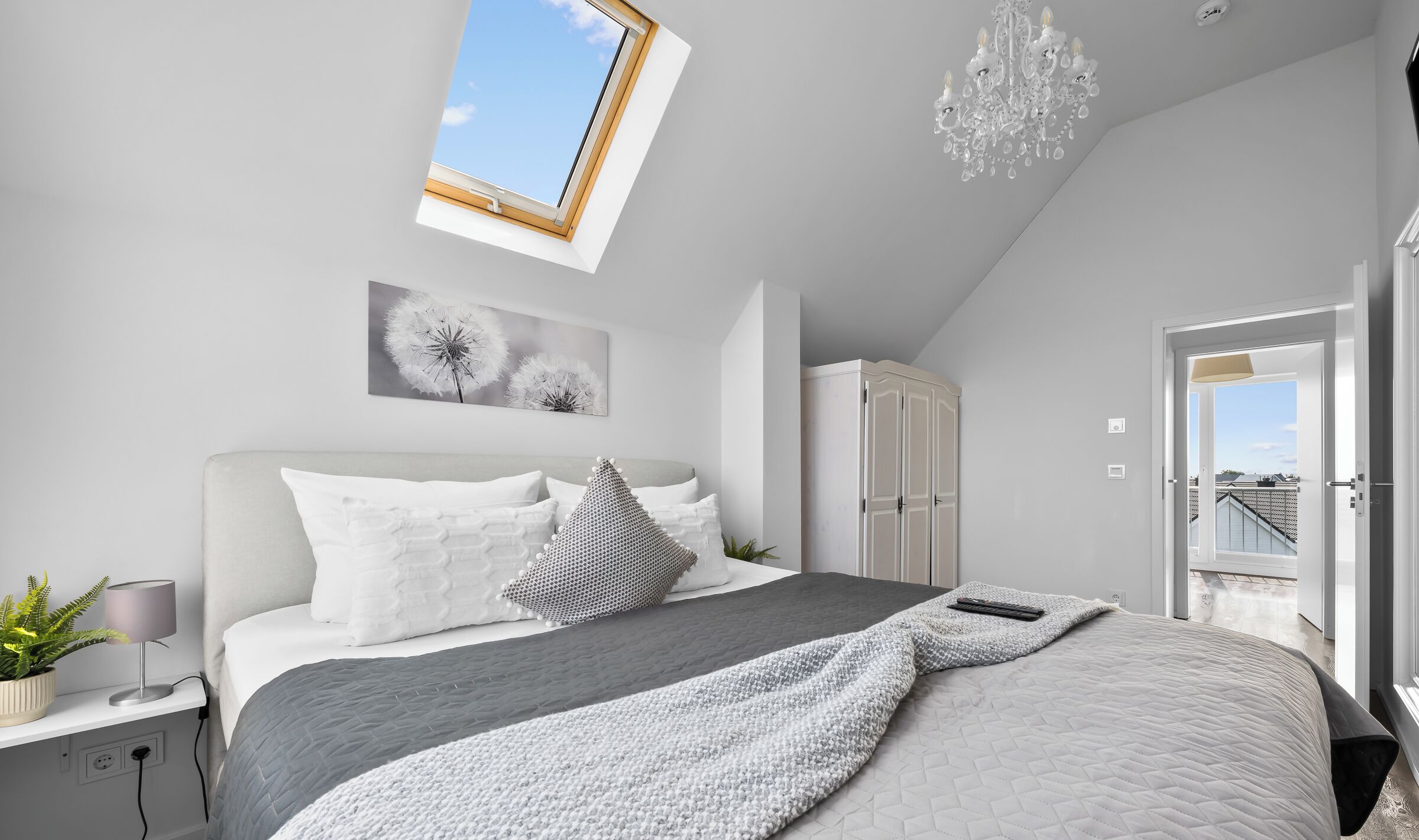 Ein modernes Schlafzimmer mit Oberlichter, einer Chandelieber, einem Bett mit vielen Kissen, einer Zimmerpflanze und einem Kleiderschrank.