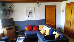 Een woonkamer met een blauwe bank, rode en gele kussens, een televisie op een houten kast en een ingelijst schilderij aan de muur.