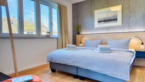 A neatly arranged bedroom with a large bed, a bedside table, a window with curtains, and a framed picture on the wall. - Mantelmöwe 32: Vacation Rental for 4 with a Balcony in the Wreecher Idyll (Putbus)