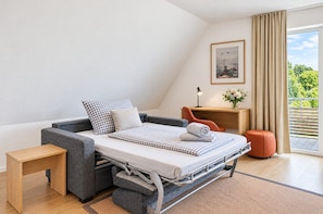Un dormitorio moderno con un sofá cama tipo futón, una mesita de noche de madera, un escritorio con lámpara, un cuadro enmarcado y vista a zonas verdes por la ventana.