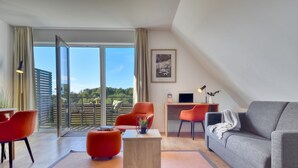 A modern living room with a grey sofa, red chairs, a wooden coffee table, and a desk with a computer. Large glass doors lead to a balcony with a view of greenery. - Mantelmöwe 32: Vacation Rental for 4 with a Balcony in the Wreecher Idyll (Putbus)
