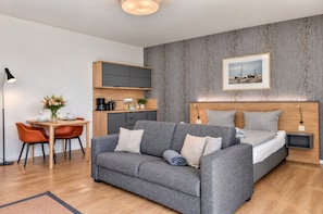 A modern hotel room with a grey sofa, a wooden headboard, a small desk with a lamp, and a framed picture on the wall. - Vacation apartment in the Wreecher Idyll on Lake Wreechen (Putbus)