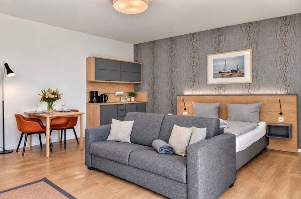 A modern hotel room with a grey sofa, a wooden headboard, a small desk with a lamp, and a framed picture on the wall.