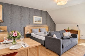 Une chambre moderne avec un lit, un canapé gris, un bureau en bois et un coin repas avec une table mise pour deux personnes.