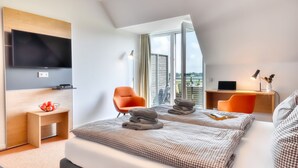 A modern hotel room with a large bed, a TV mounted on the wall, a desk with a laptop, and a view of the city through large windows. - Vacation apartment in the Wreecher Idyll on Lake Wreechen (Putbus)