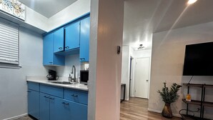 Eine kleine Küche mit blauen Schränken, einem Spülbecken und einer Arbeitsplatte. Rechts befindet sich ein kleiner Fernseher und eine Pflanze.