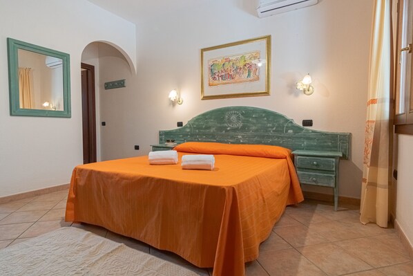 A bedroom with a green headboard, an orange bedspread, two white towels, a nightstand, a dresser, a framed picture, and a mirror with a green frame.