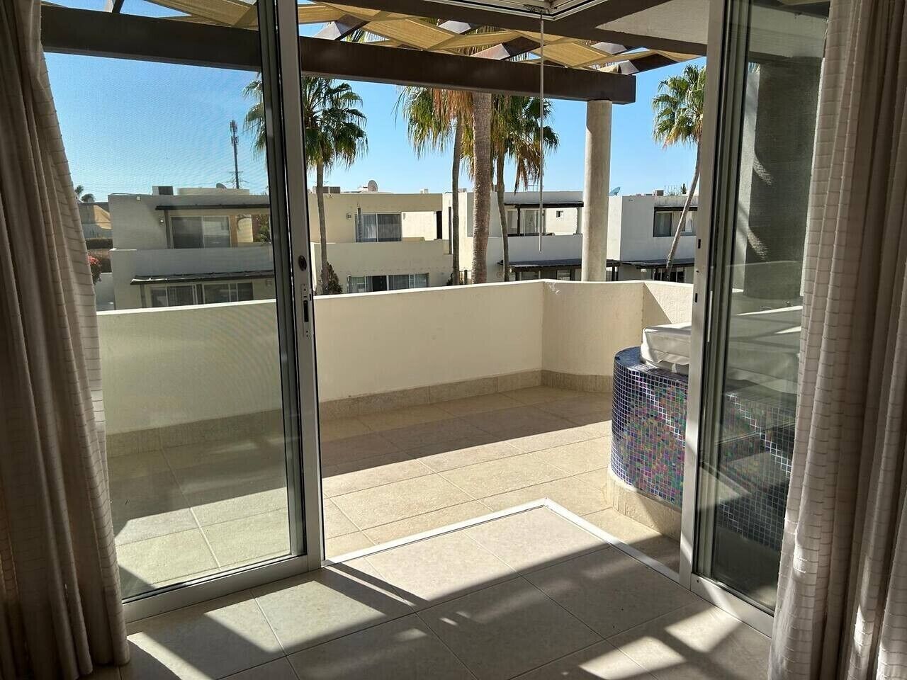 Een balkon met uitzicht op palmbomen en gebouwen, een betegelde vloer en een glazen deur.
