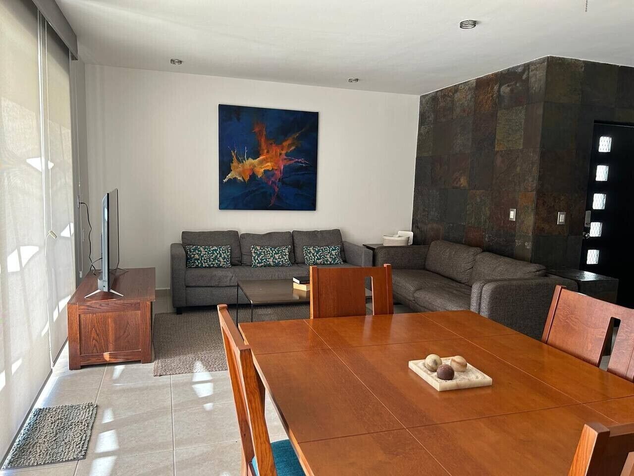 Een moderne woonkamer met een houten eettafel, grijze banken en een groot abstract schilderij aan de muur.