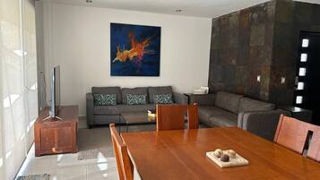 Een moderne woonkamer met een houten eettafel, grijze banken en een groot abstract schilderij aan de muur.