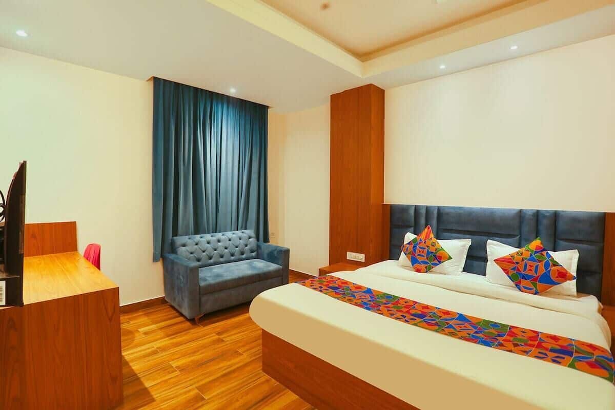 A hotel room with a wooden desk, a blue sofa, a bed with colorful pillows, and a large window with blue curtains.