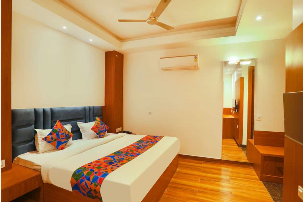 A hotel room with a bed, a ceiling fan, an air conditioning unit, wooden flooring, and a small seating area.