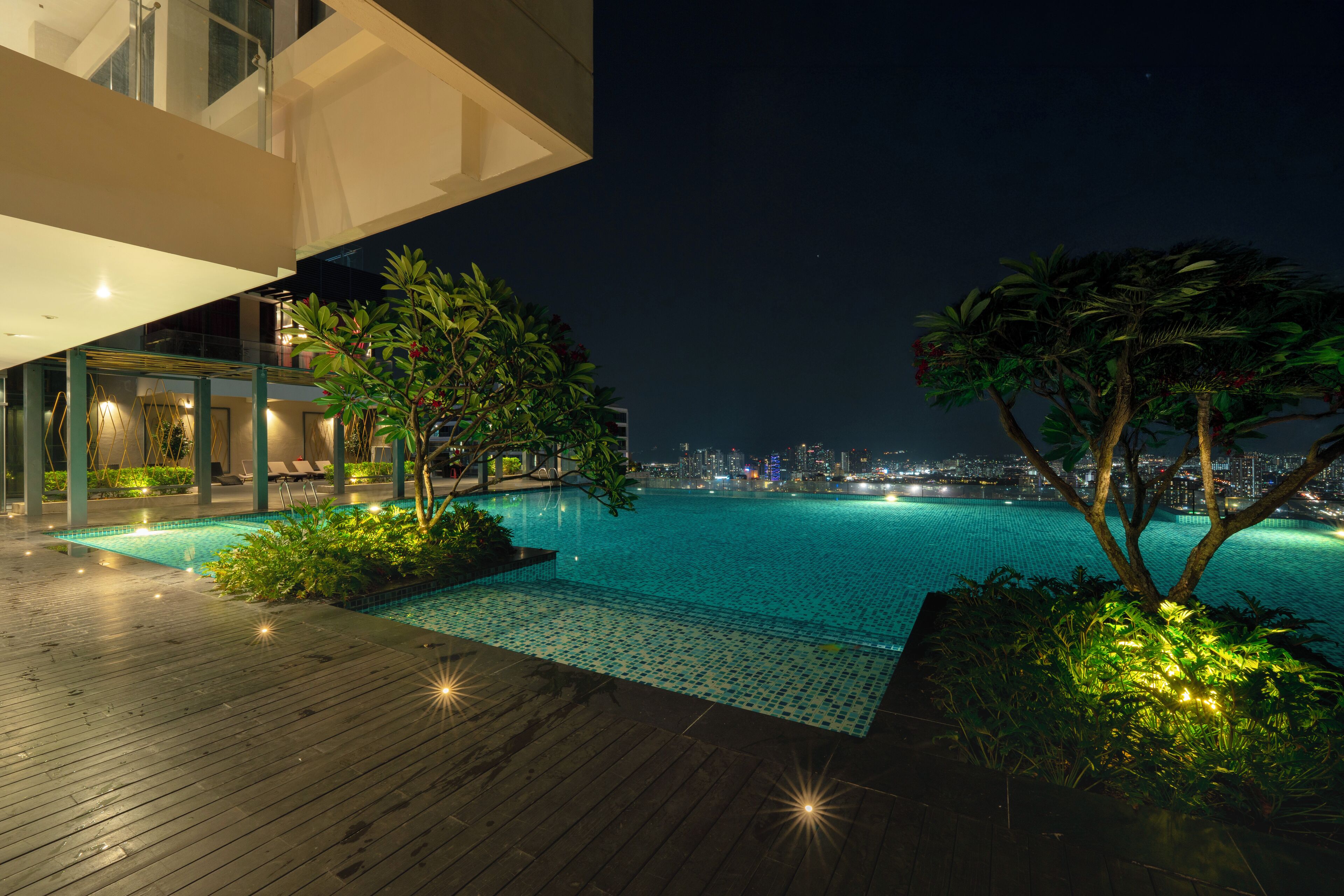 Un espace piscine extérieur moderne, doté d’une terrasse en bois, entouré d’une végétation luxuriante et offrant une vue sur les toits de la ville la nuit.