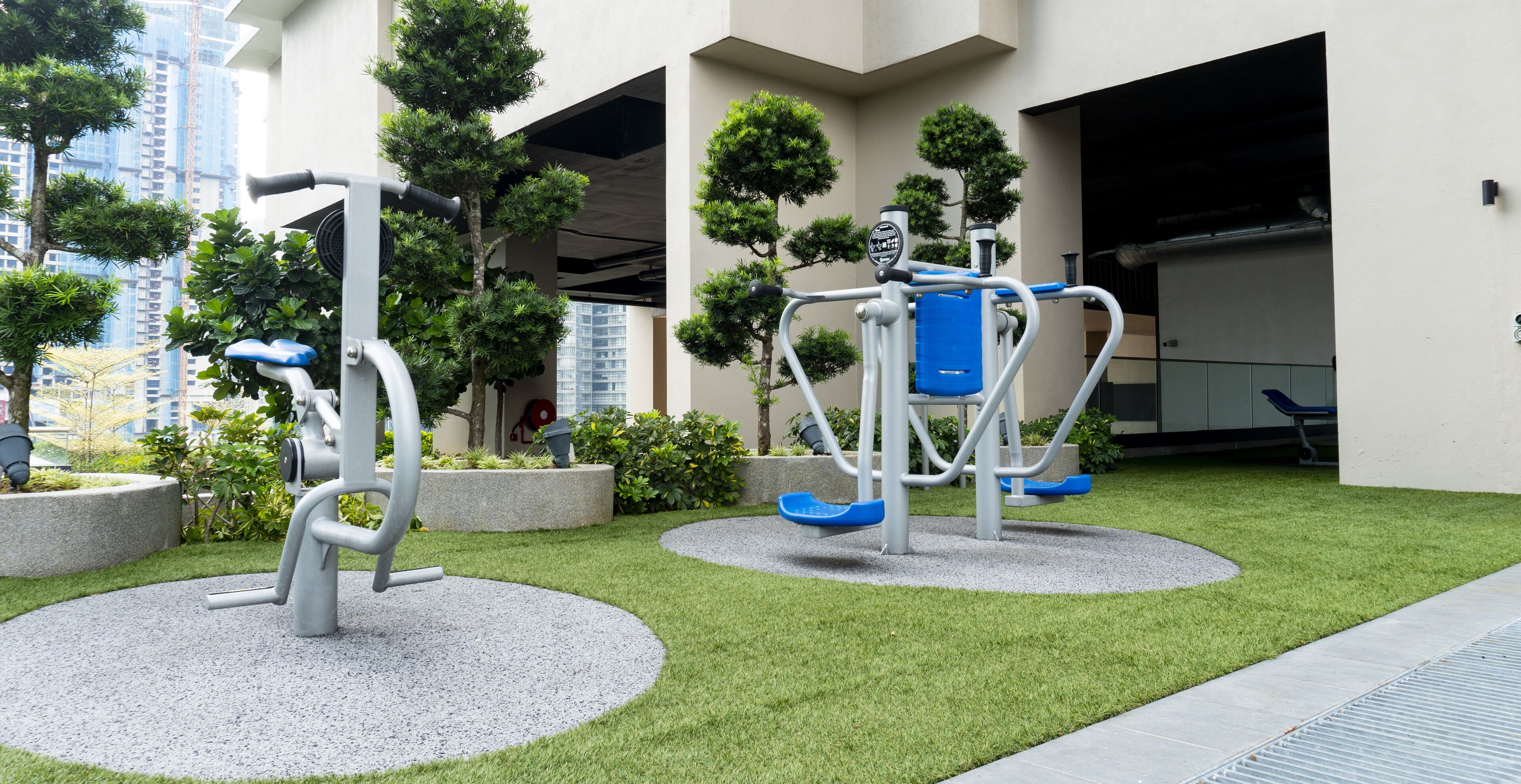 Outdoor exercise equipment in a courtyard with artificial grass, surrounded by planters and a building in the background.