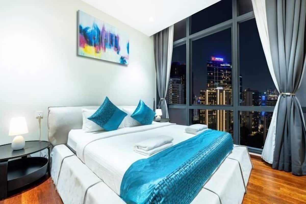 Quarto de hotel moderno com uma grande janela, cama com roupa de cama branca e detalhes em azul, mesa de cabeceira e vista para a cidade.