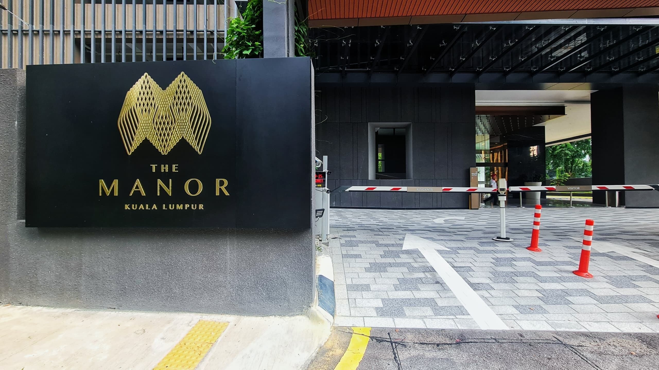 A hotel entrance with a black sign displaying 'The Manor Kuala Lumpur' and a gated entrance with red and white striped barriers.