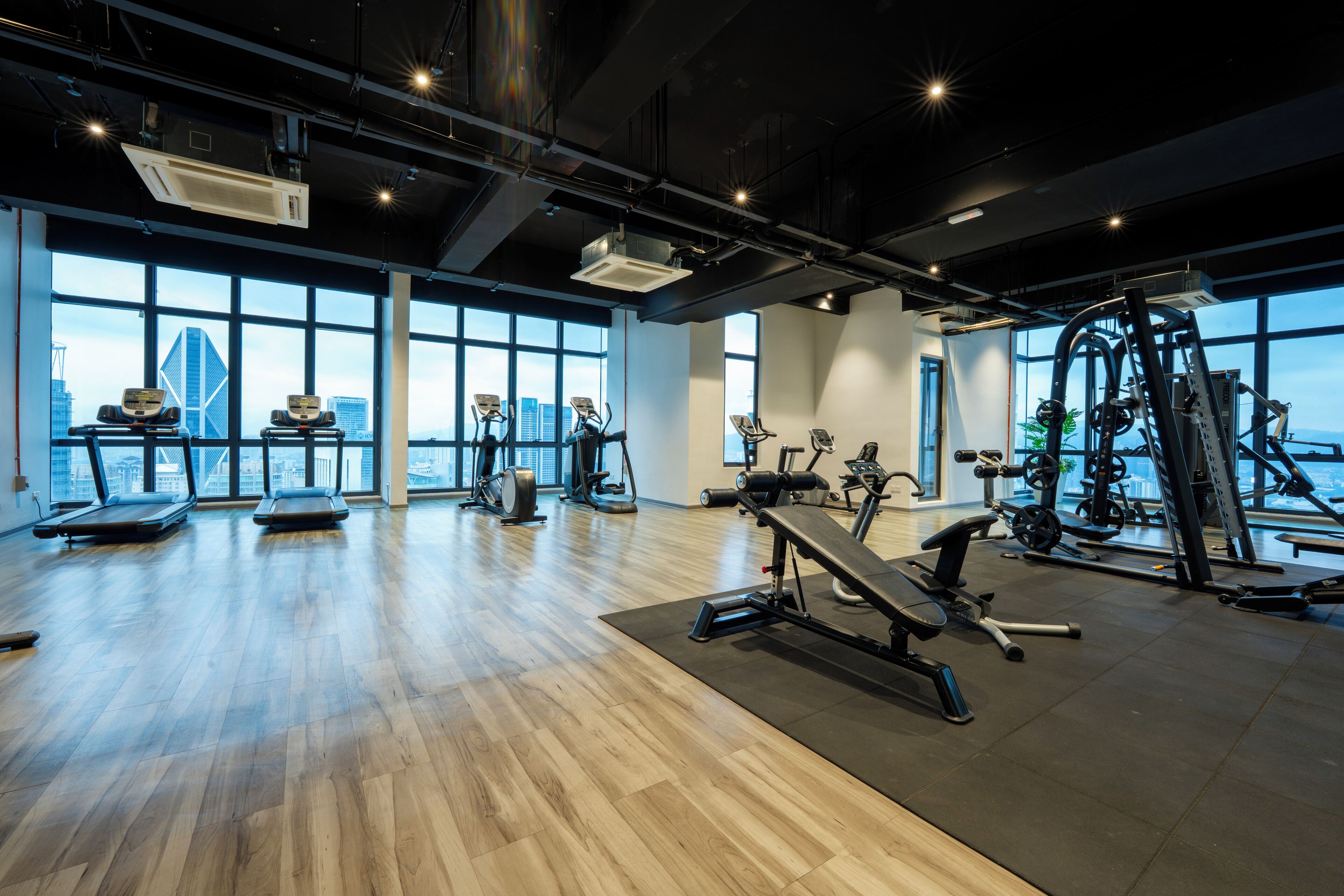 A spacious gym with various exercise equipment, large windows offering a city view, and a polished wooden floor.