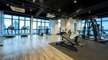 A spacious gym with various exercise equipment, large windows offering a city view, and a polished wooden floor.