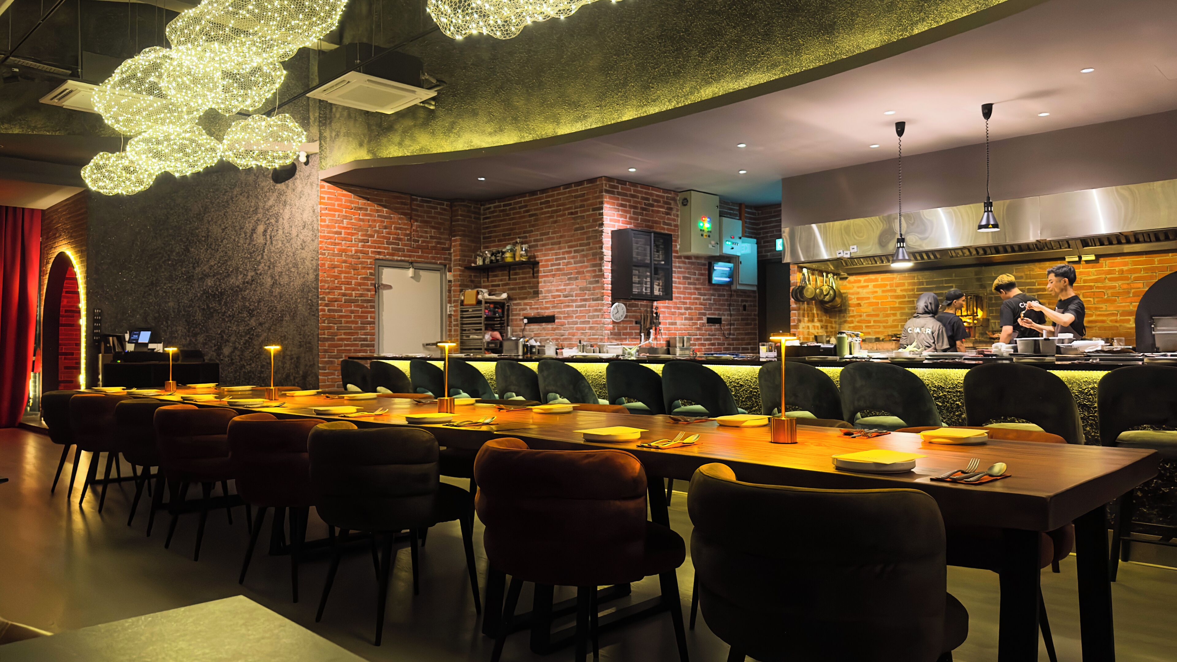 A restaurant with a long communal table, individual booths, a brick wall, and a kitchen area visible in the background.