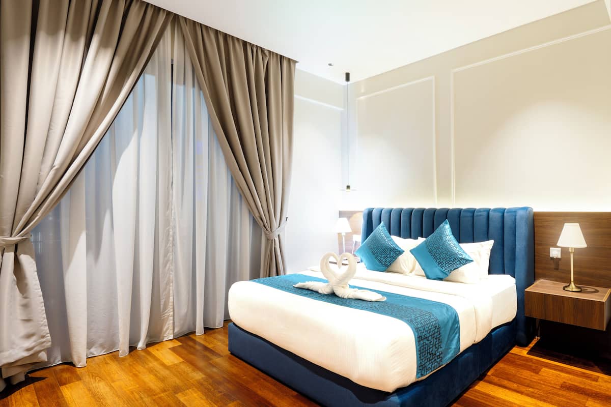 Quarto de hotel com uma cama grande, cabeceira, mesa de cabeceira e janela com cortinas.