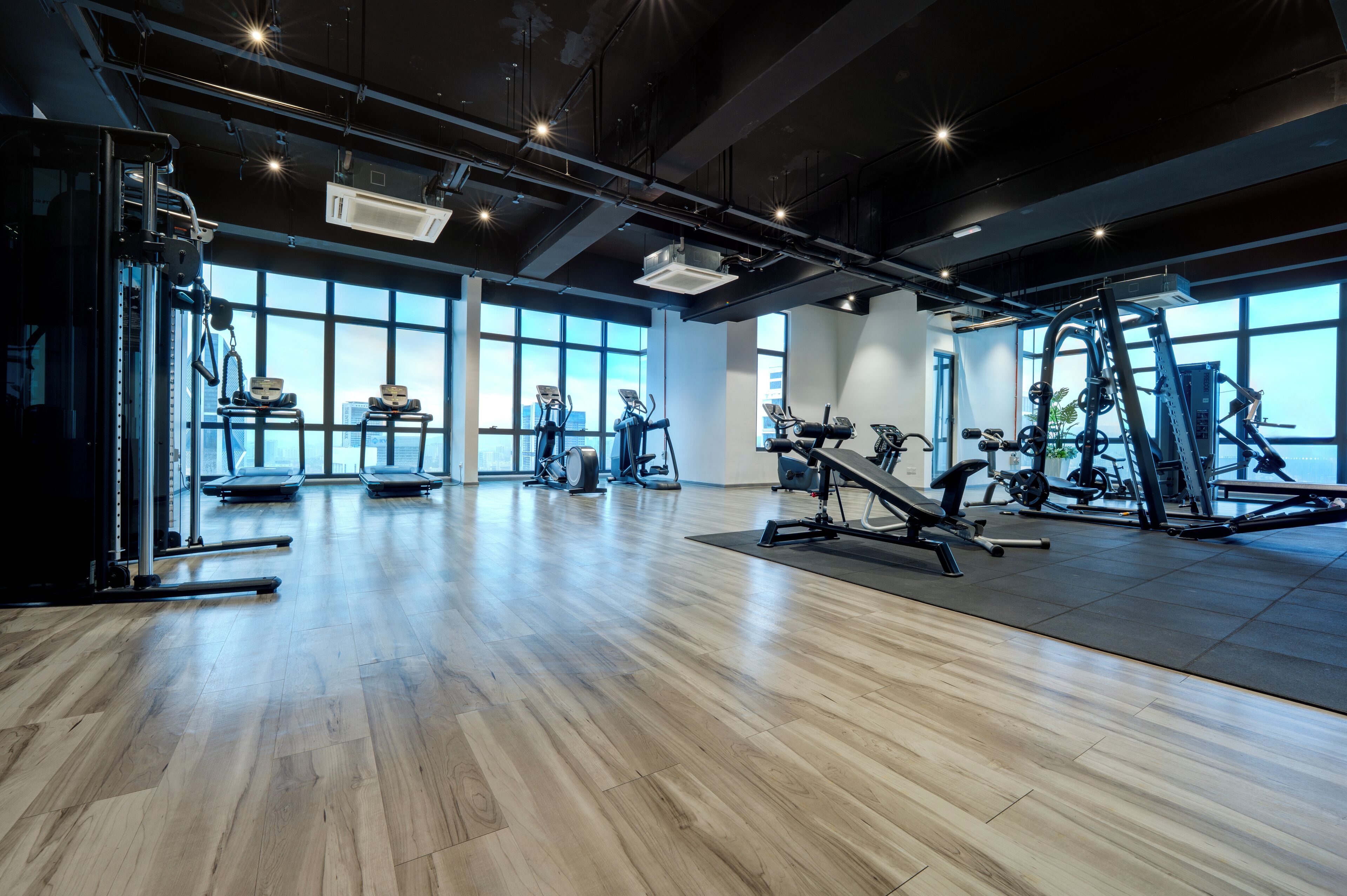 A spacious gym with various exercise equipment, large windows, and a polished wooden floor.