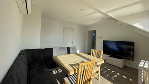 Un salon moderne avec un canapé d’angle noir, une table à manger en bois avec des chaises, un téléviseur à écran plat et une lucarne.