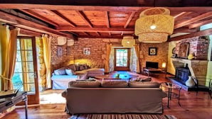 A rustic cabin interior with a stone wall, wooden ceiling beams, a sofa, a fireplace, and a hanging light fixture. - La Fermette au Banquet, Les Eyzies (Les Eyzies)
