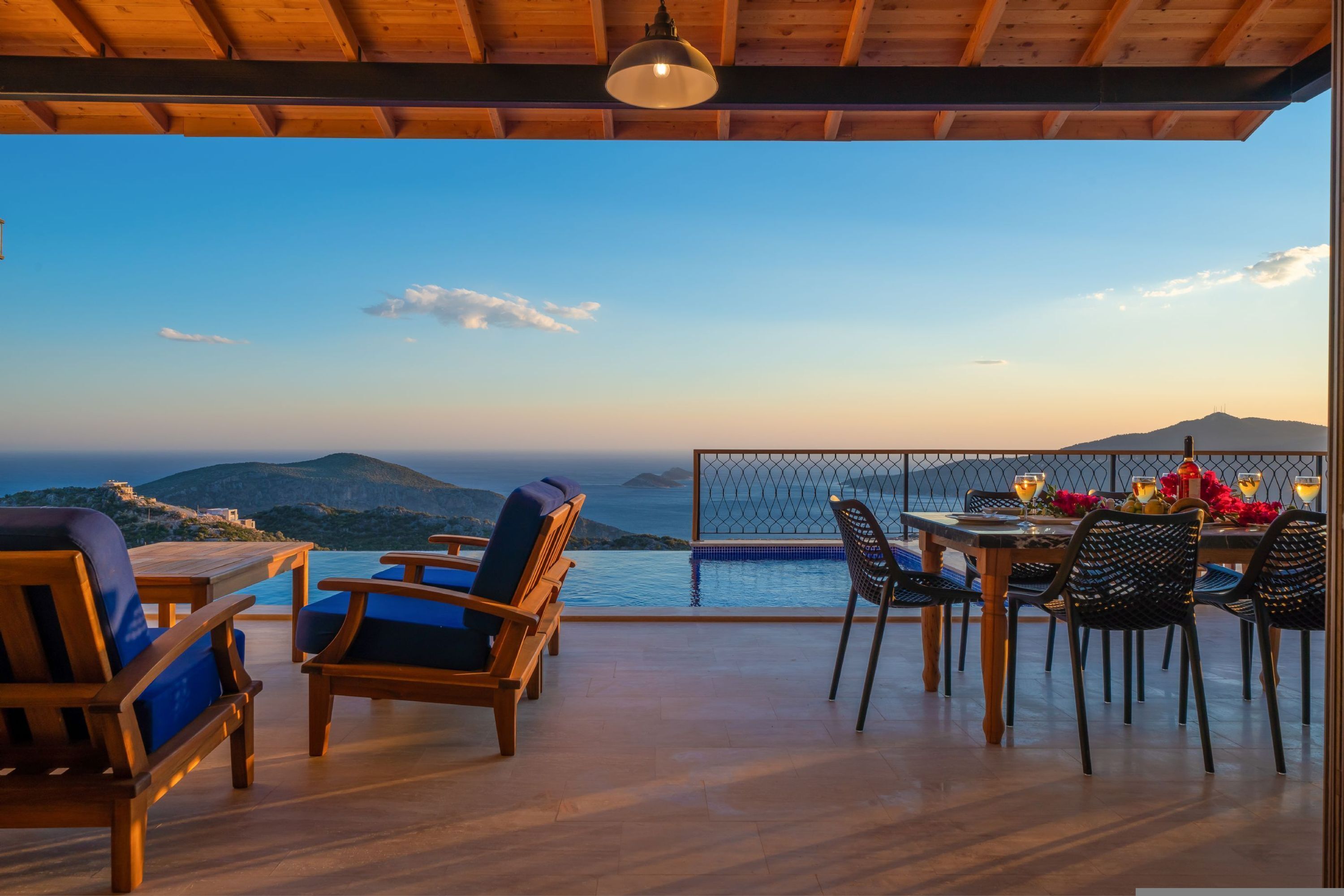 A terrace with wooden furniture, a dining table set with chairs, and a view of the sea and mountains.