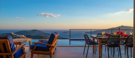 A terrace with wooden furniture, a dining table set with chairs, and a view of the sea and mountains.