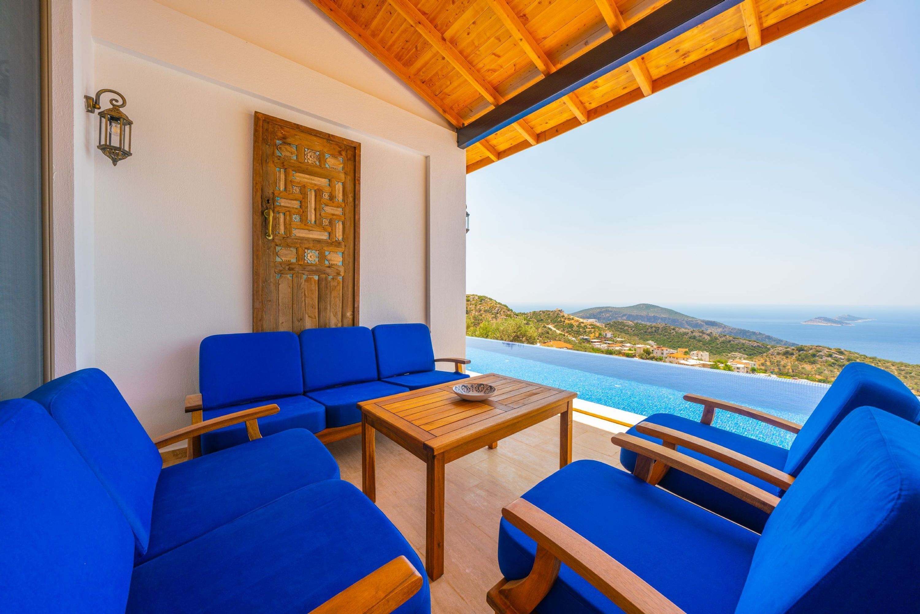 Une terrasse avec des canapés bleus et une table en bois, offrant une vue sur une piscine et un paysage pittoresque de la mer et des collines.