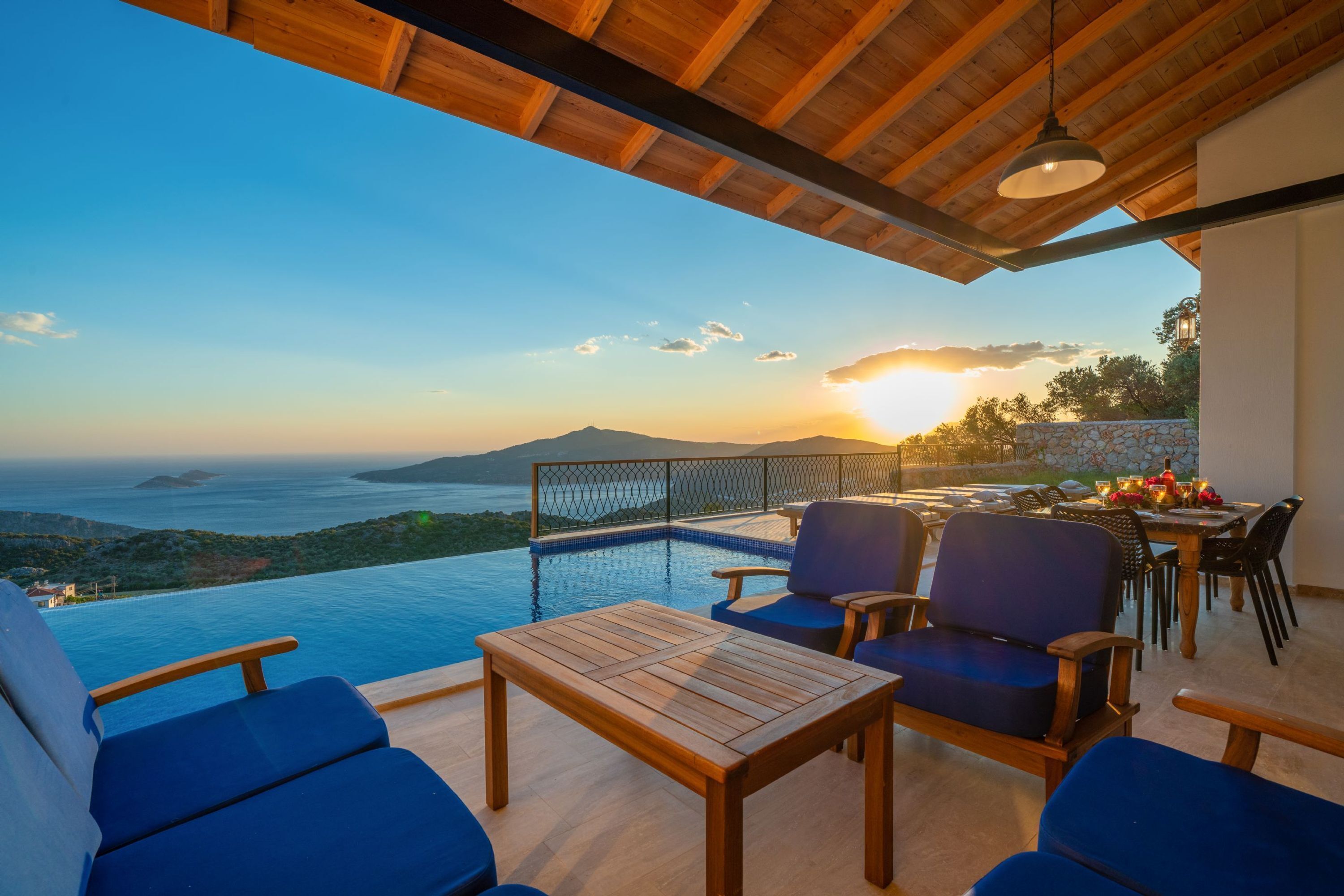 A terrace with a wooden table and chairs, overlooking a pool and a scenic view of the sea and mountains.