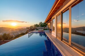 Ein moderner Infinity-Pool mit Blick auf den Sonnenuntergang über den Bergen, eine Holzterrasse und große Glasfenster.