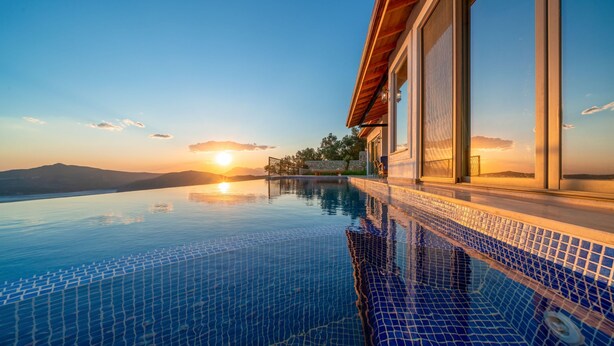 En modern infinitypool med fri utsikt över solnedgången över bergen, en stenmur och ett trädäck.