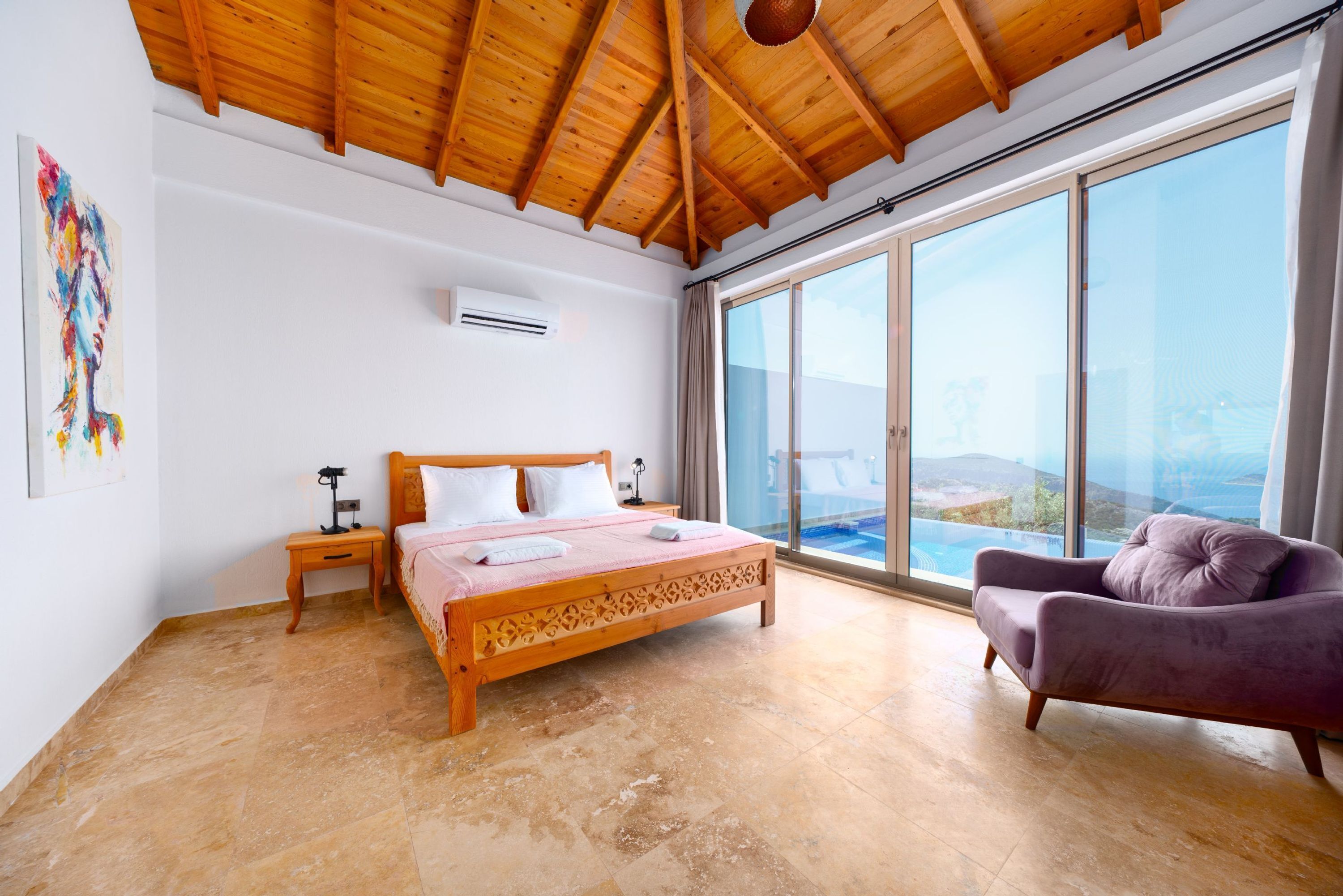 Une chambre avec un plafond en bois, un grand lit, un fauteuil violet et une vue sur la mer grâce à de grandes fenêtres.