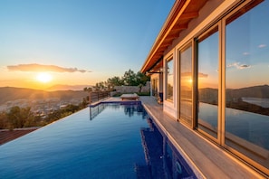 Uma piscina infinita moderna com vista para o pôr do sol sobre as montanhas, um deck de madeira e grandes portas de vidro.