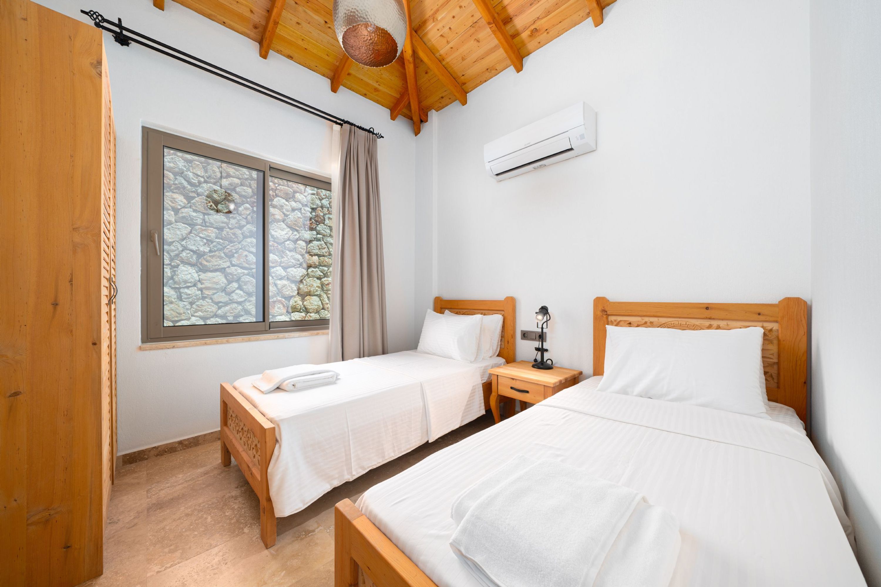 A bedroom with two beds, a window with stone walls, an air conditioner, and wooden ceiling beams.