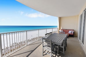 A balcony with a table and chairs, a sofa with cushions, and a view of the beach and ocean.