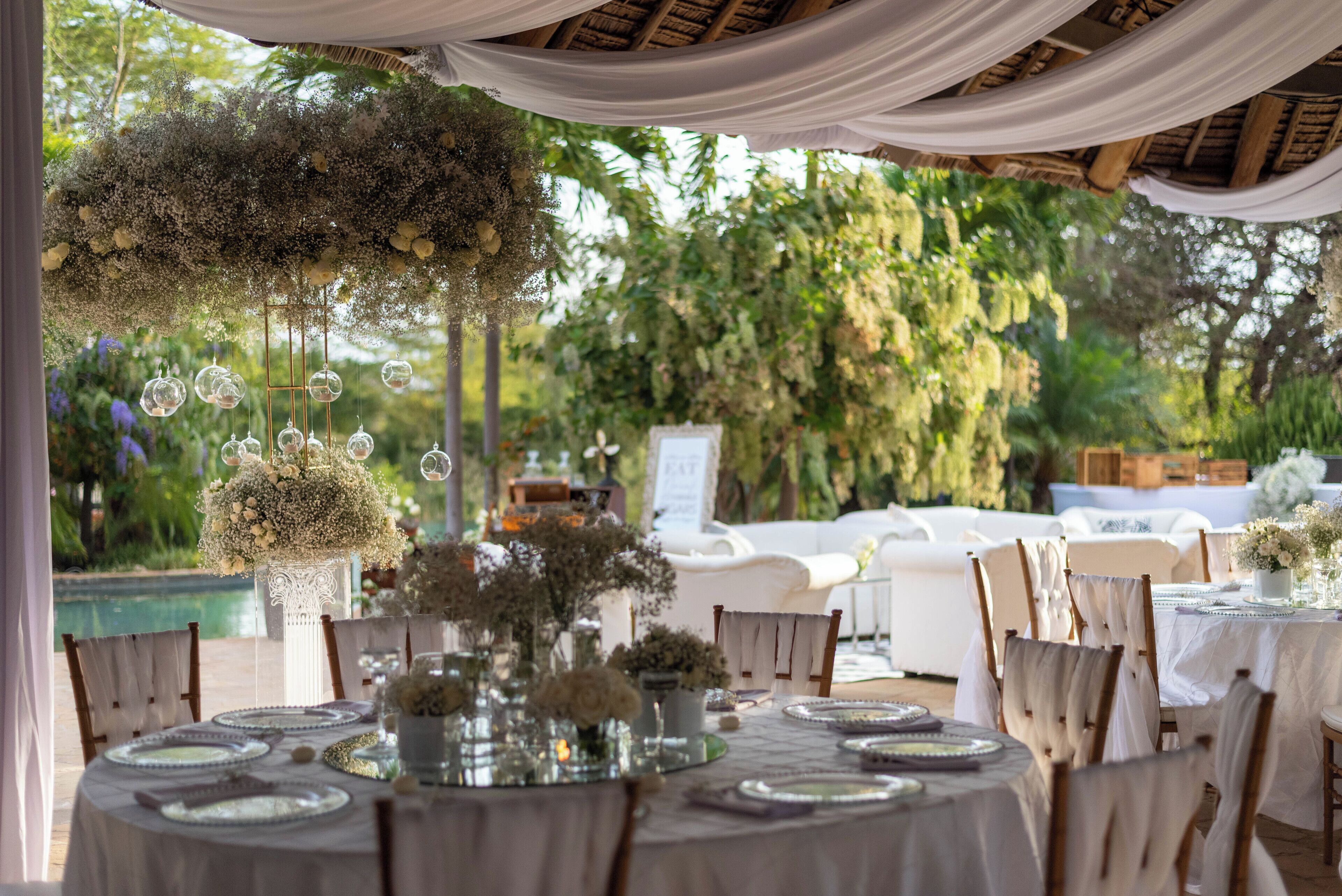 Un salón de bodas al aire libre con mesas redondas dispuestas para cenar, rodeado de sillas blancas y cubiertas con manteles blancos.