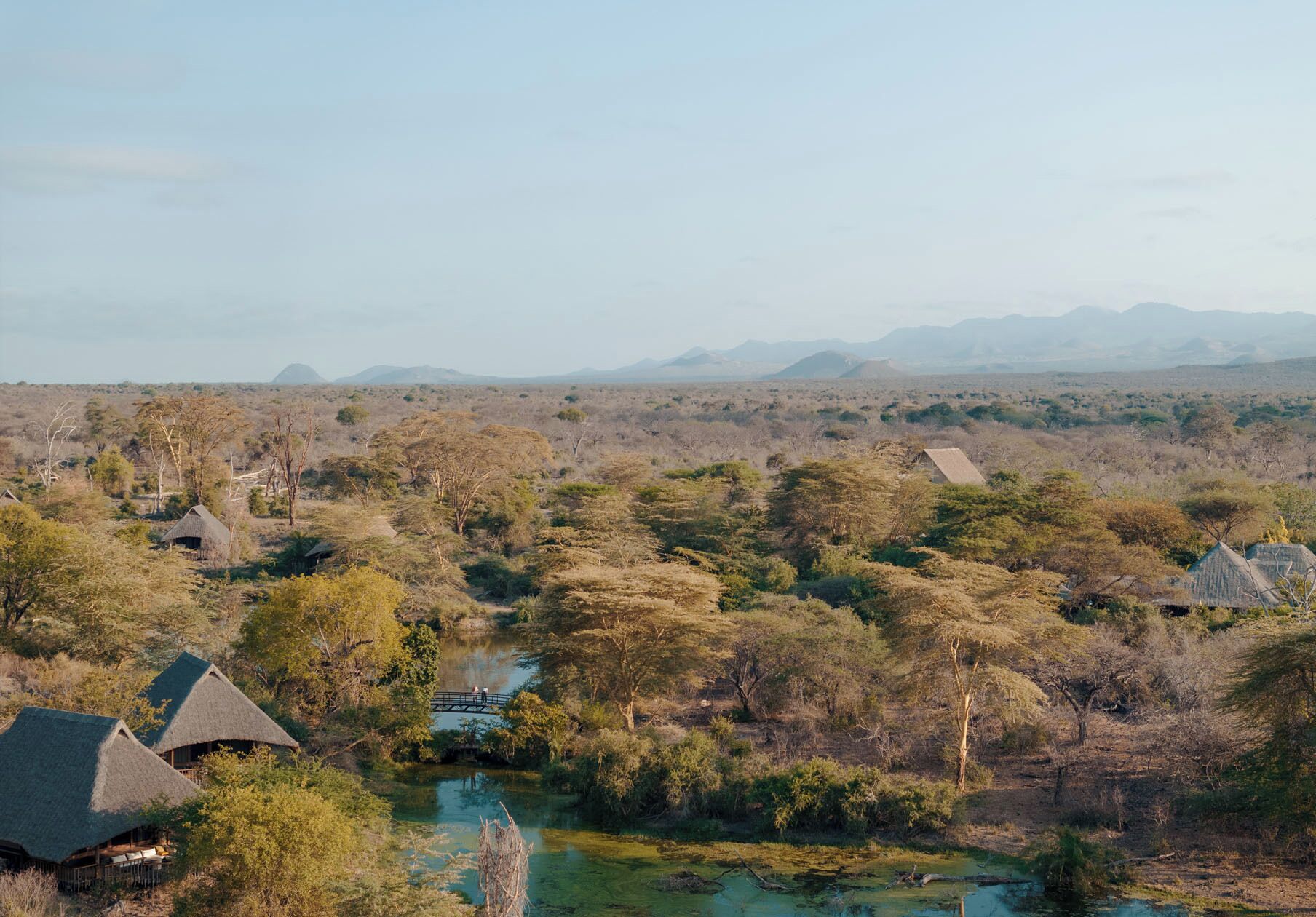 Un lodge de safari con construcciones de techo de paja, rodeado por un paisaje de sabana con árboles dispersos y una cadena montañosa distante.