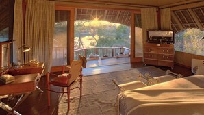 A spacious bedroom with a large bed, a desk with a lamp, a chair, a TV, and a balcony with a view of the outdoors. - Finch Hattons (Tsavo)